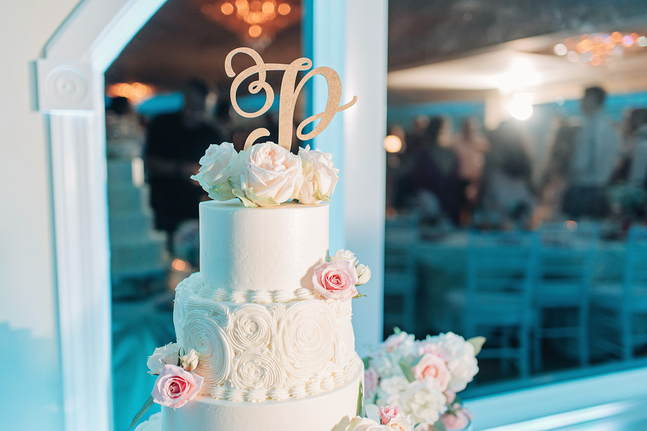 Elegant three-tier wedding cake adorned with roses and monogram topper at reception.