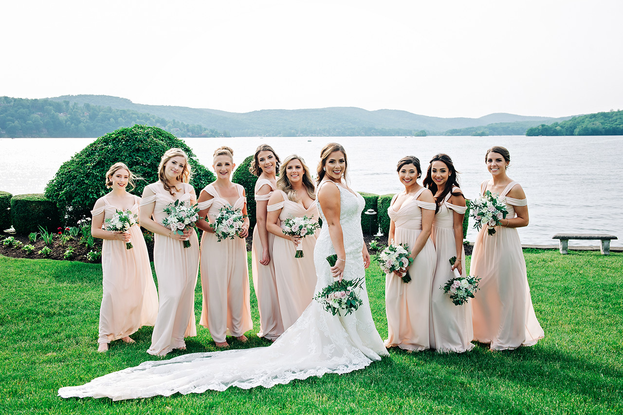 Bride and bridesmaids pose with bouquets by a scenic lakeside wedding venue.
