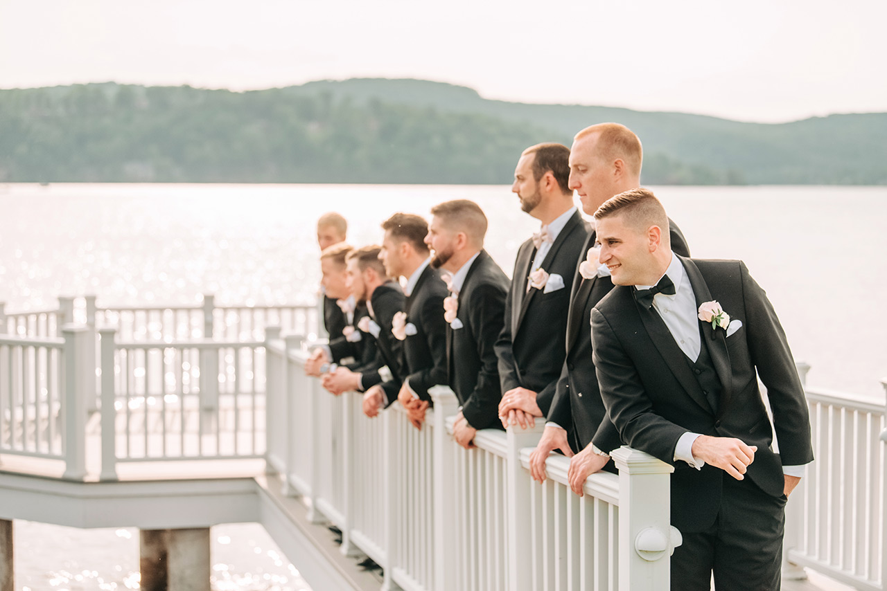 Groomsmen in formal attire posing on a lakeside pier during a waterfront wedding celebration.