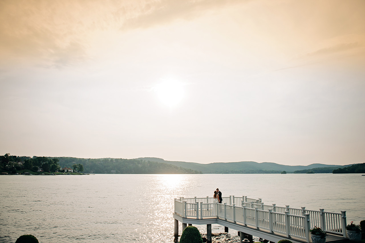 Romantic couple embracing on a lakeside pier at sunset with shimmering water and scenic views.