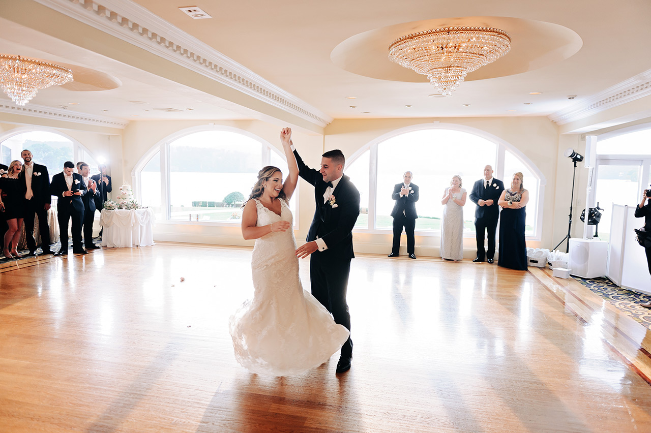 Bride and groom share their first dance in a bright ballroom as guests look on.