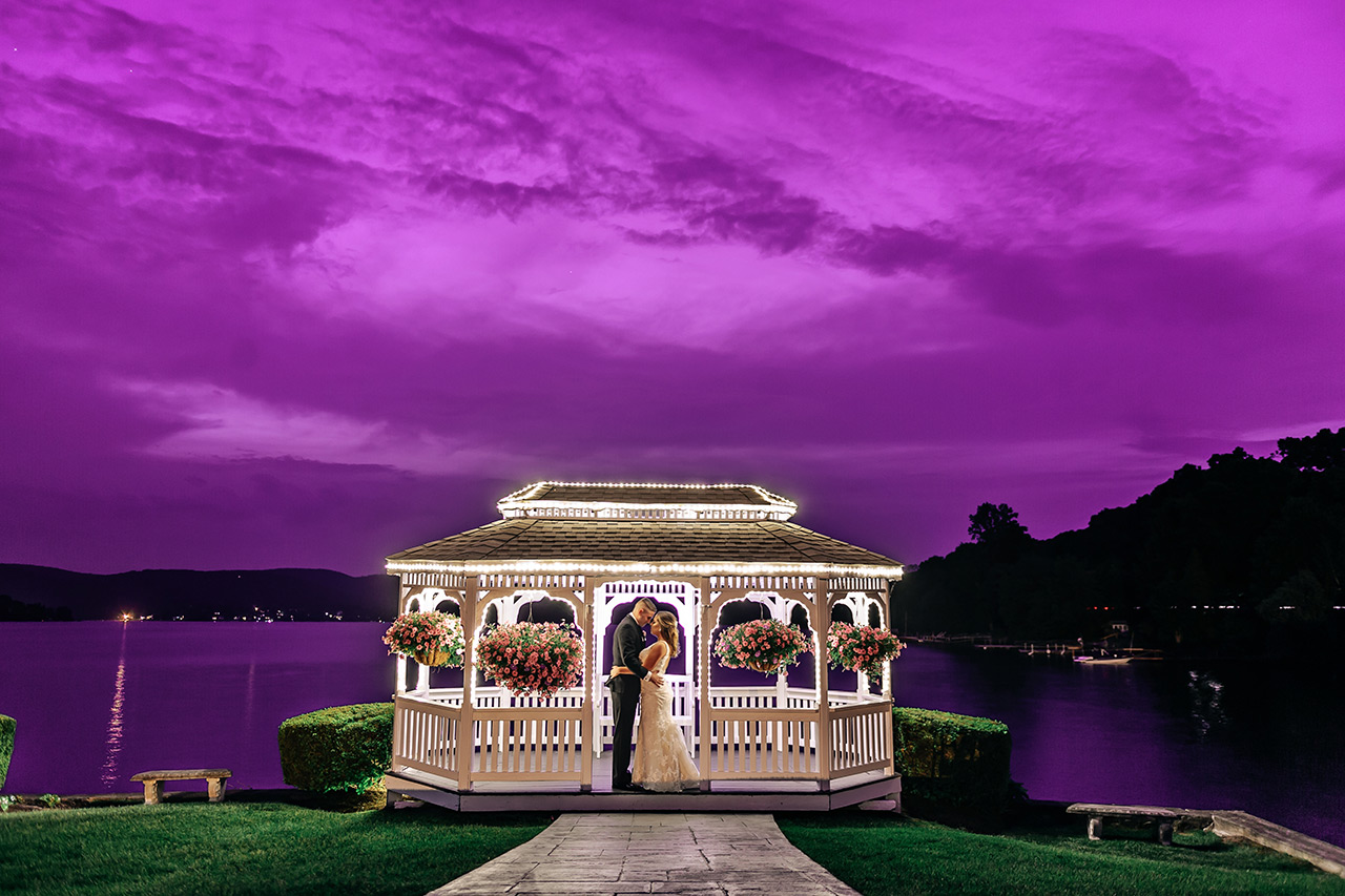Bride and groom embrace in a lakeside gazebo beneath a dramatic purple evening sky.