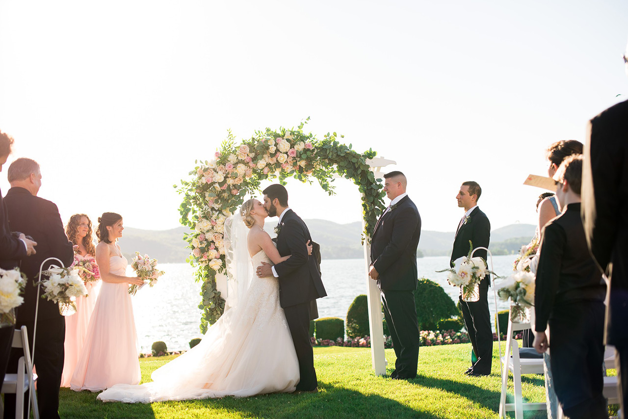 Bride and groom share a kiss beneath a floral arch during a lakeside wedding ceremony.