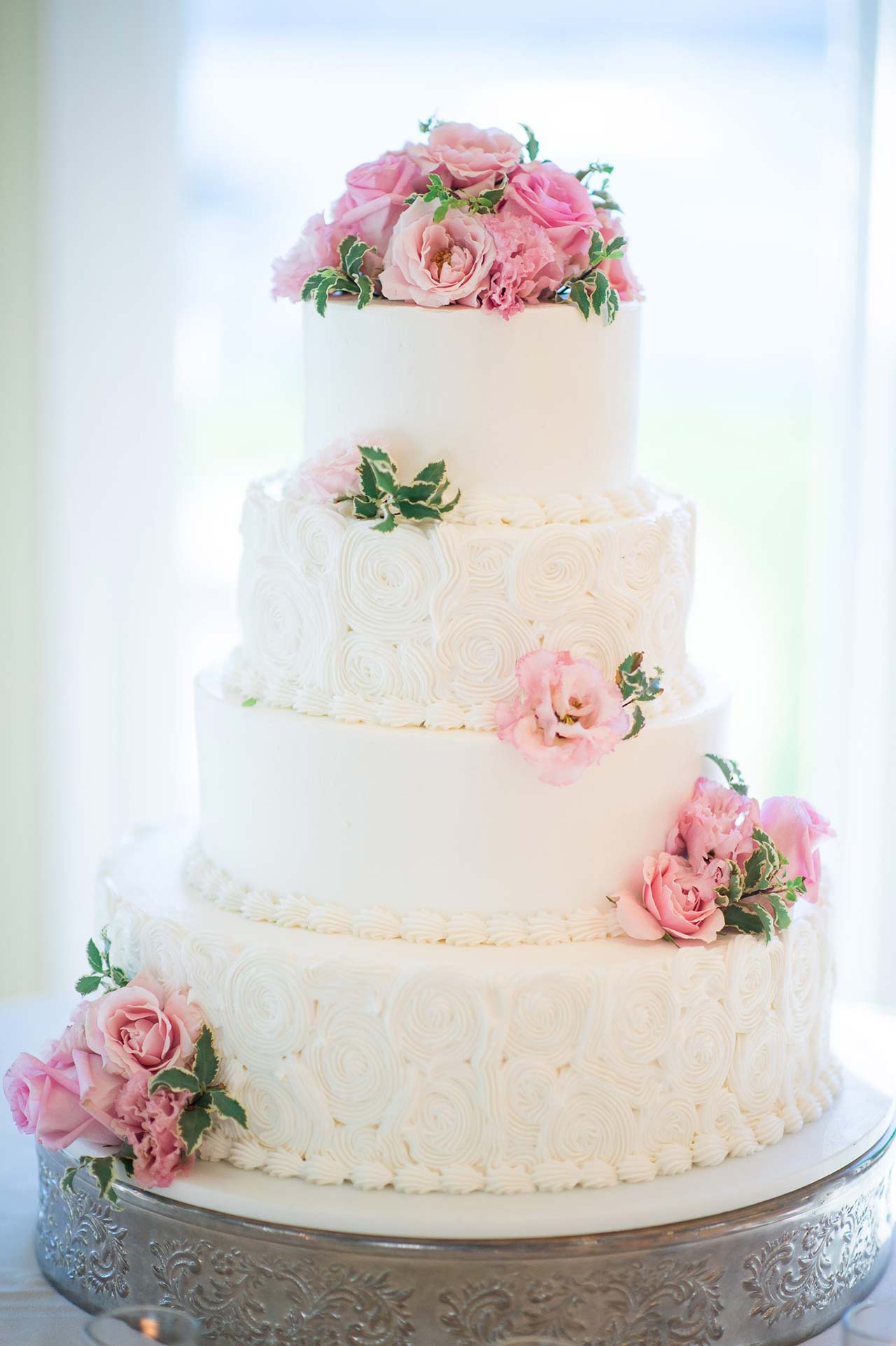 Four-tier white wedding cake decorated with pink roses and elegant buttercream swirl detailing.