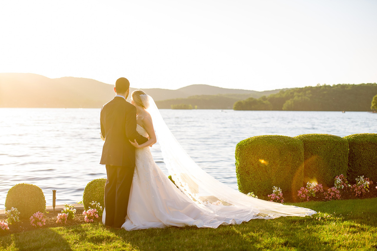 Bride and groom embrace by the lake at sunset, with flowing veil and scenic views.
