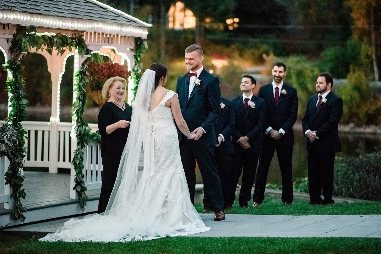 Bride and groom exchange vows beside a lakeside gazebo during an elegant outdoor wedding ceremony.