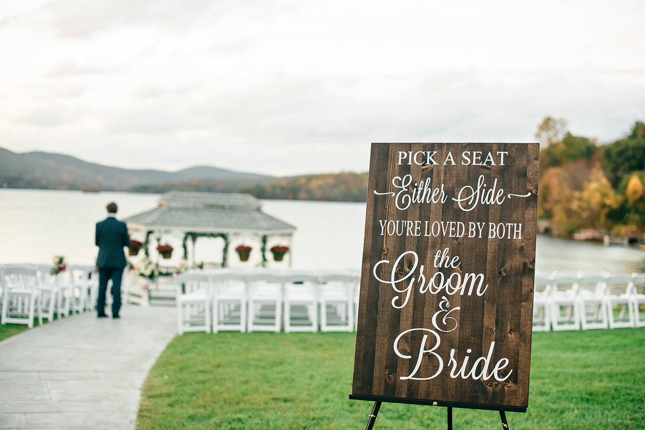 Rustic wedding welcome sign beside a lakeside ceremony setup with gazebo and seating.