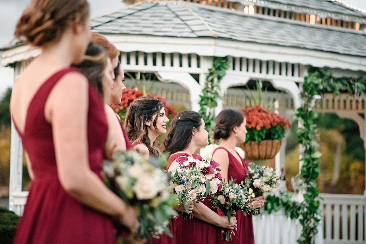 Bridesmaids in burgundy dresses hold bouquets beside a decorated gazebo during an outdoor wedding.