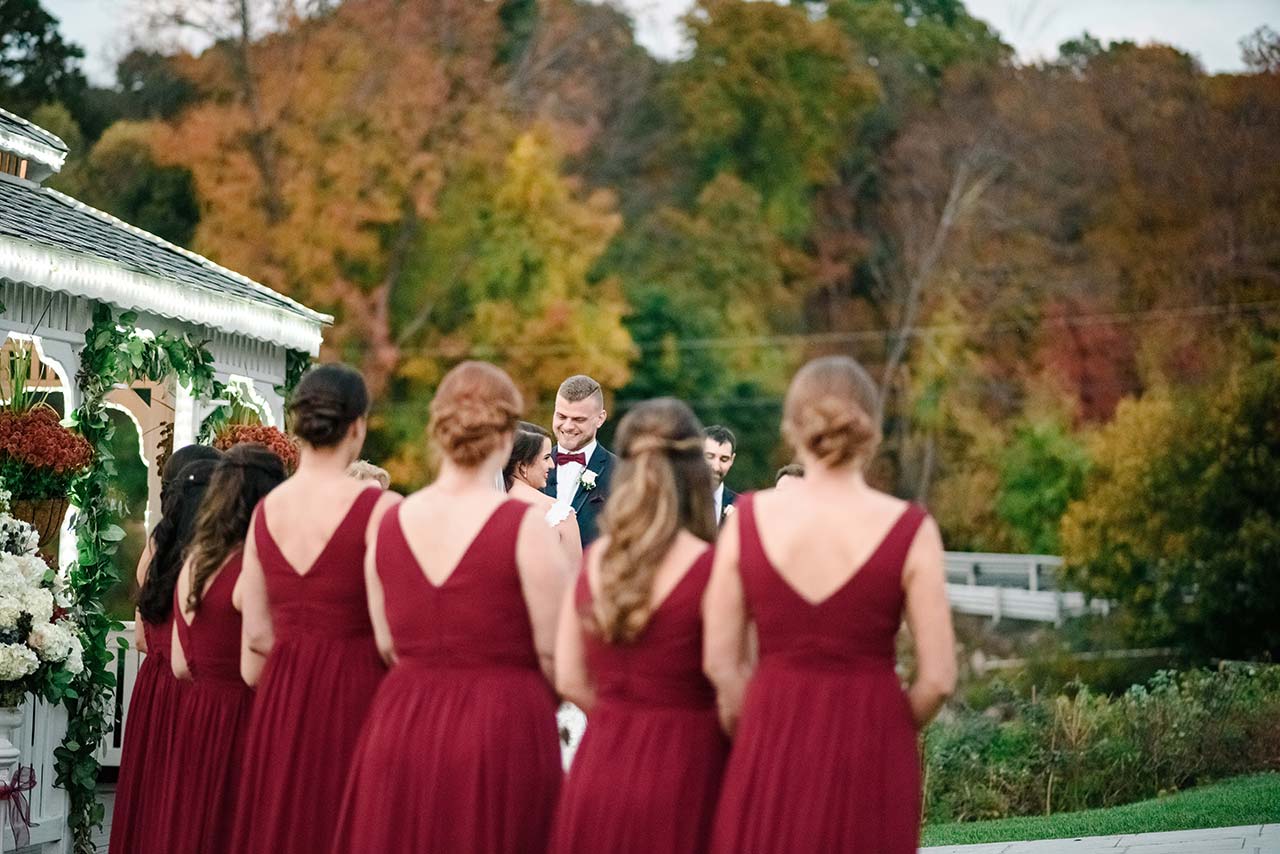 Bridesmaids in burgundy dresses watch an outdoor wedding ceremony beside a decorated gazebo.