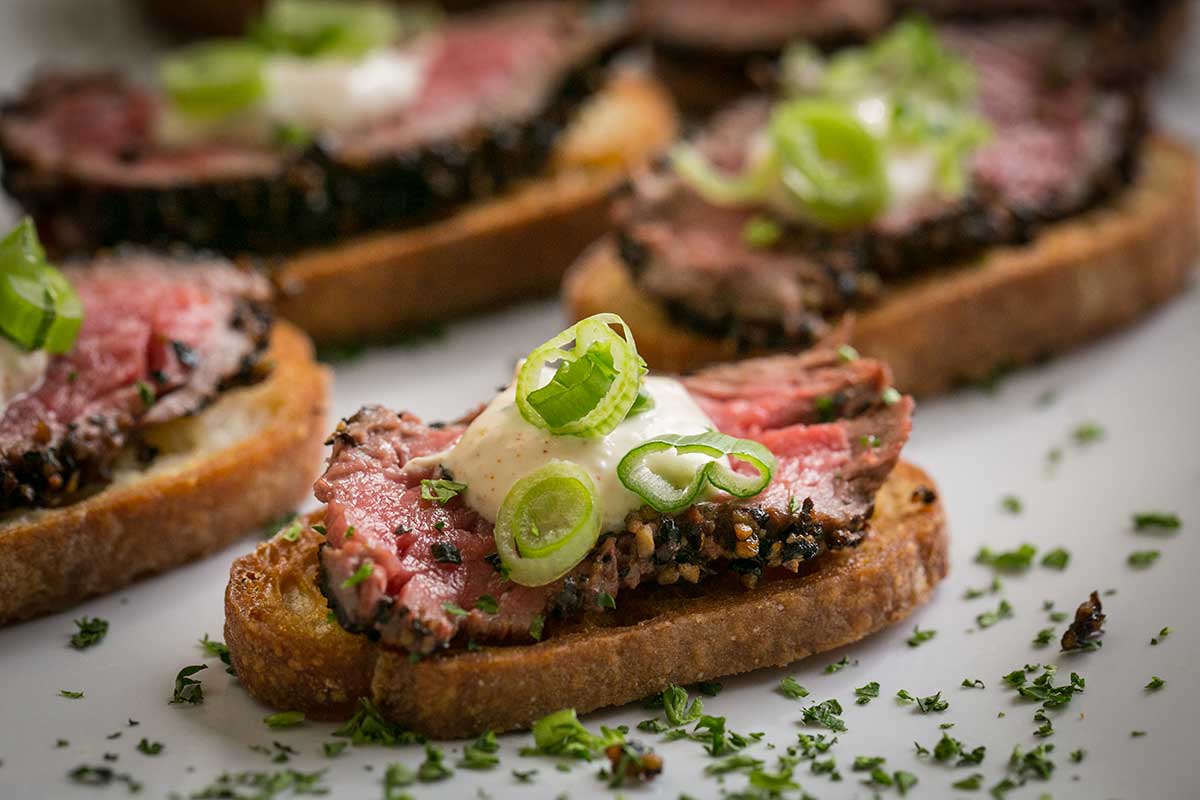 Pepper-crusted sirloin crostini topped with creamy sauce, sliced scallions, and fresh herbs.