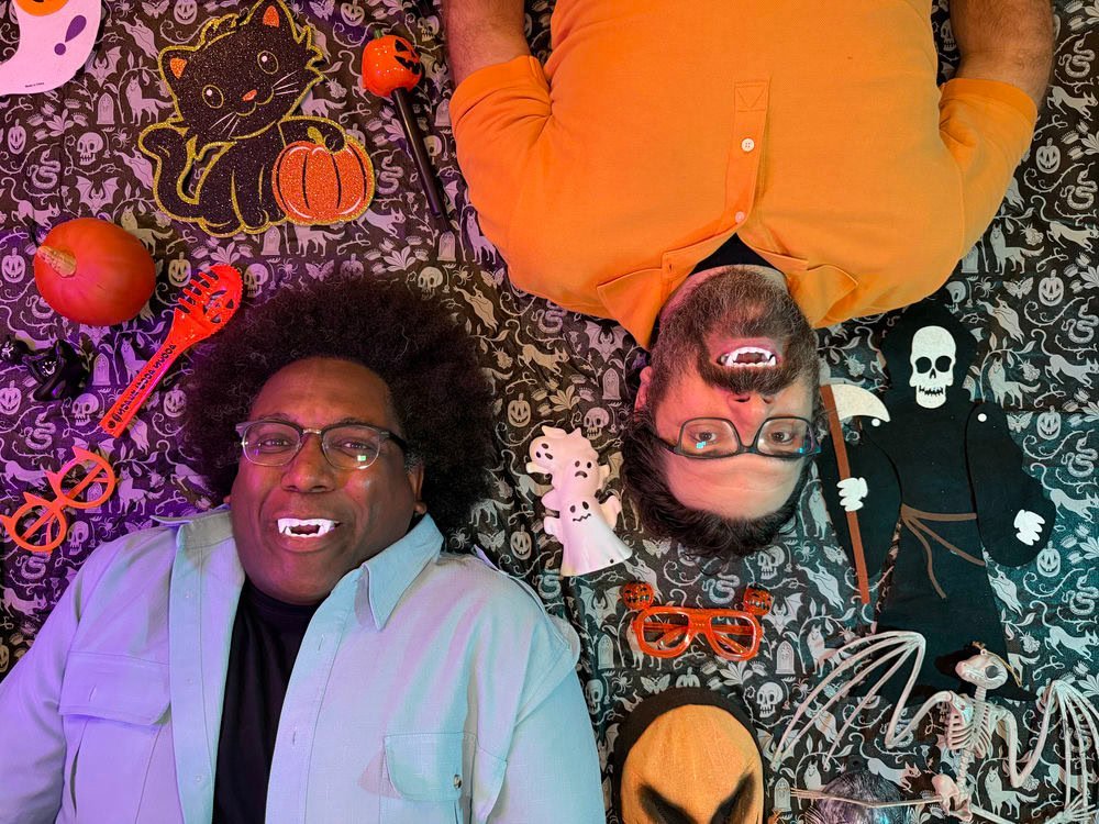 Two men lying on a Halloween-themed floor surrounded by spooky decorations at SpookyFest in Hollywood, CA.