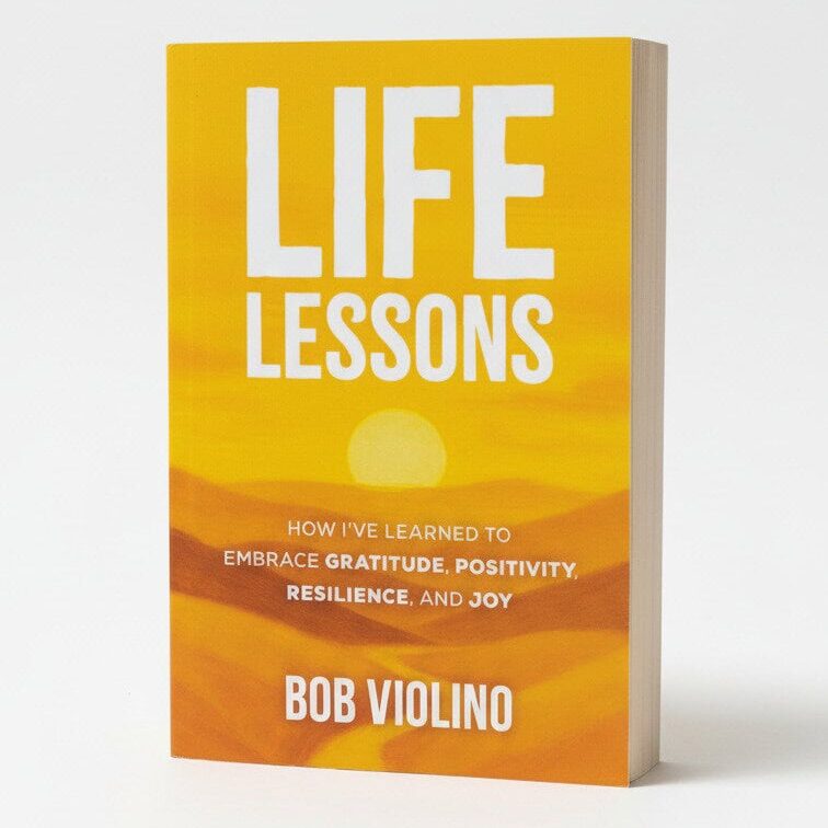 A book titled "Life Lessons" by Bob Violino stands upright against a white background. The yellow-orange cover, featuring a sun and hills, highlights resilience strategies, positivity, and joy learned through embracing gratitude.
