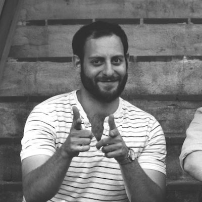 A black and white photo of a smiling man in a striped shirt, sitting and making a peace sign with both hands towards the camera, symbolizing Generation Freedom.