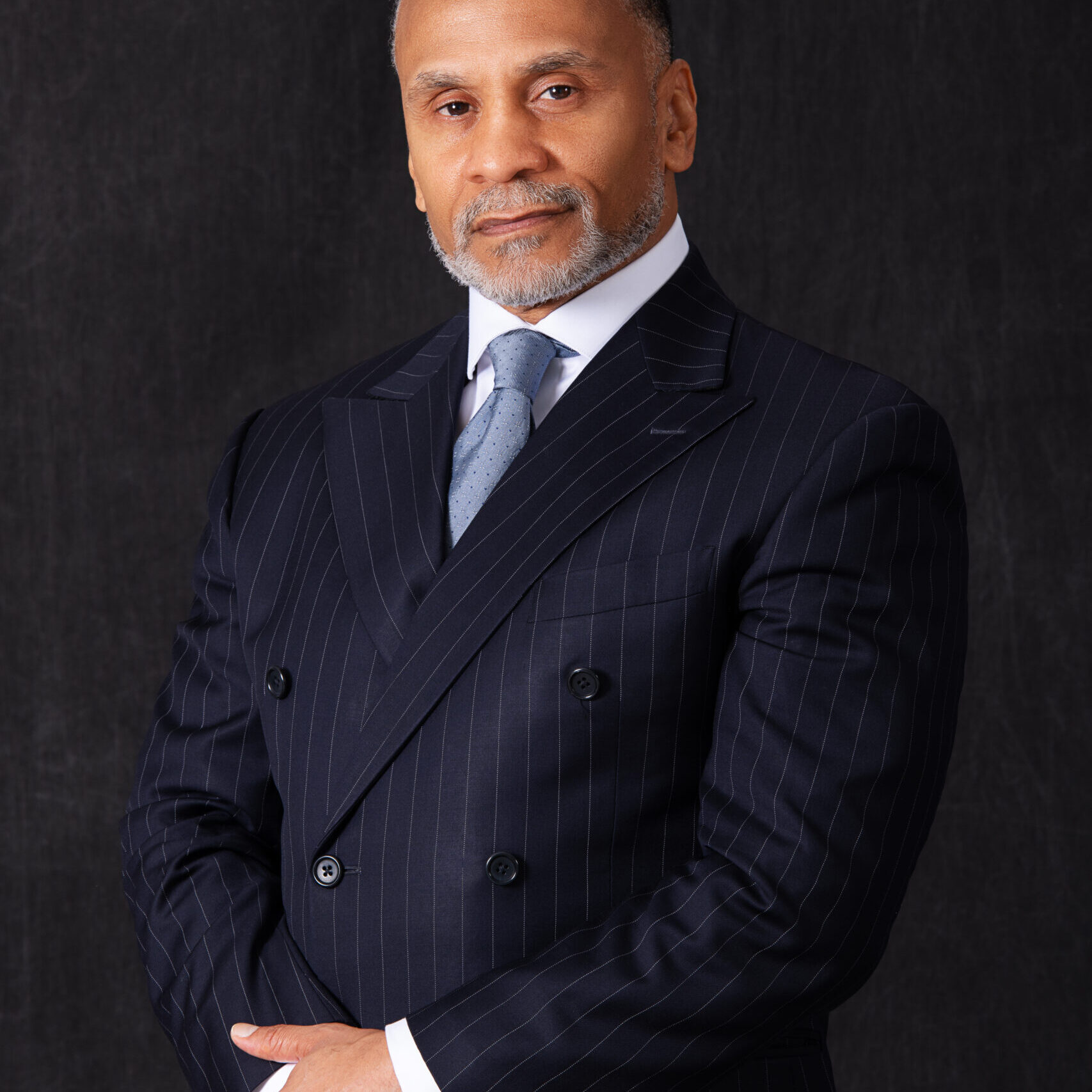 A man, Ted Santos, in a dark pinstripe suit with a light blue tie stands against a plain background, looking confidently at the camera with his hands clasped—embodying the spirit of breakthrough initiatives in corporate culture.