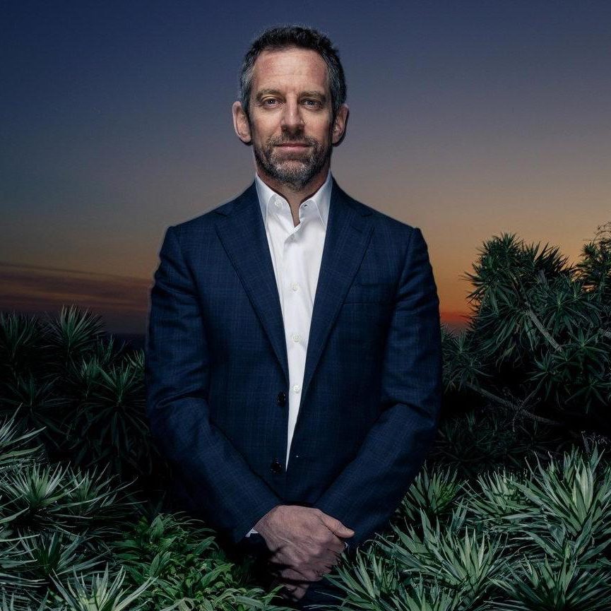 A man in a dark suit stands outdoors at dusk, reminiscent of the contemplative tone found in book reviews. Surrounded by greenery and beneath a twilight sky that transitions from blue to orange, the scene evokes themes of choice and freewill, much like Sam Harris's insightful discussions.