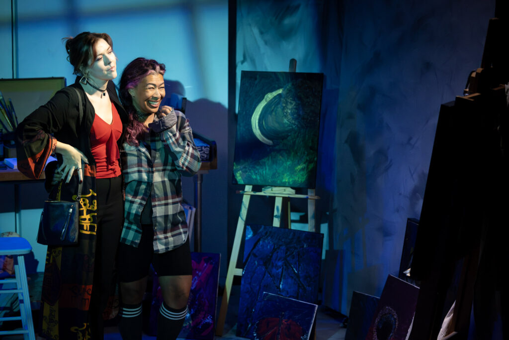 Two women in a theatrical production representing an exhibition, admiring paintings with a dark, moody atmosphere and vibrant blue lighting, showcasing creative artwork.
