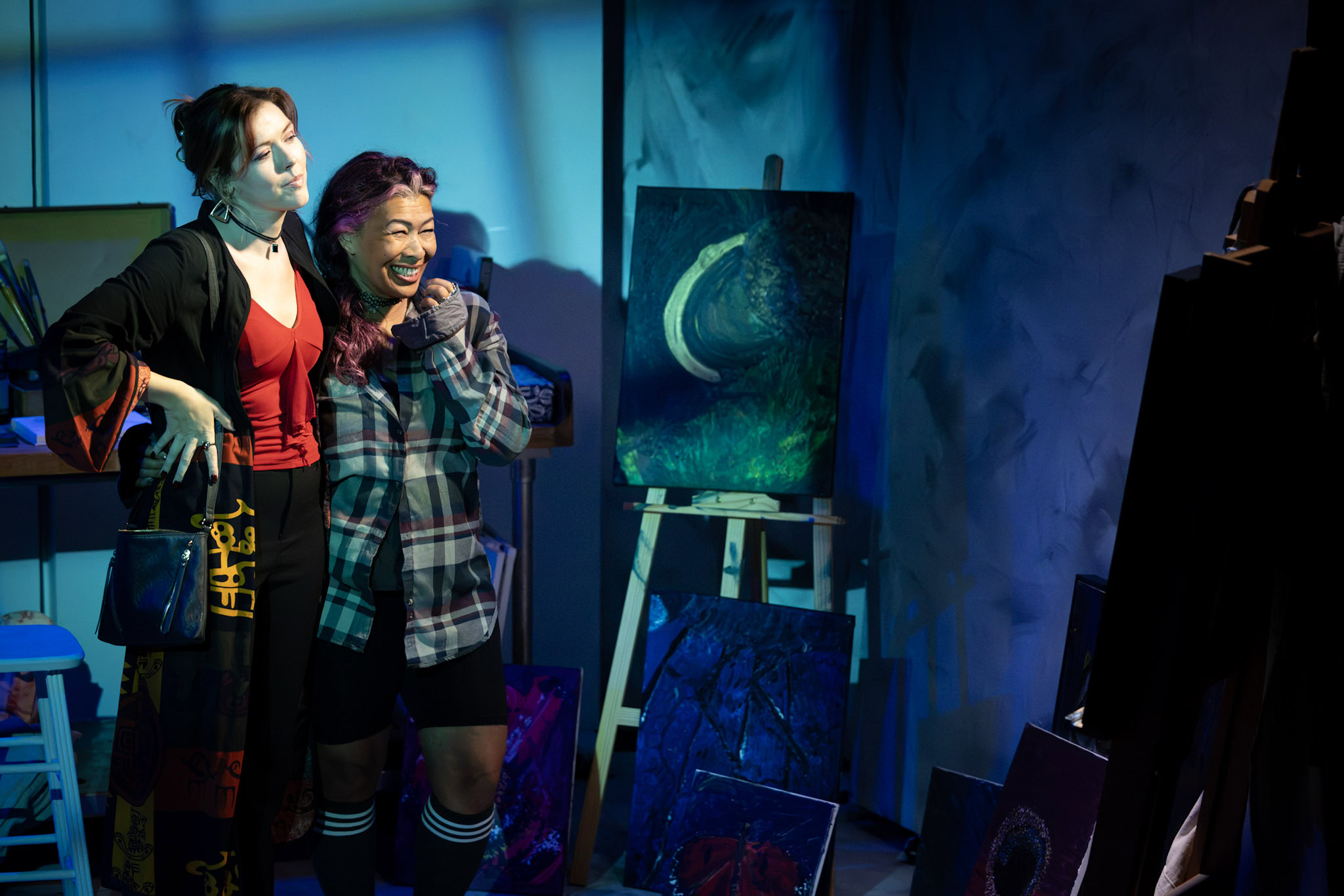 Two women in a theatrical production representing an exhibition, admiring paintings with a dark, moody atmosphere and vibrant blue lighting, showcasing creative artwork.