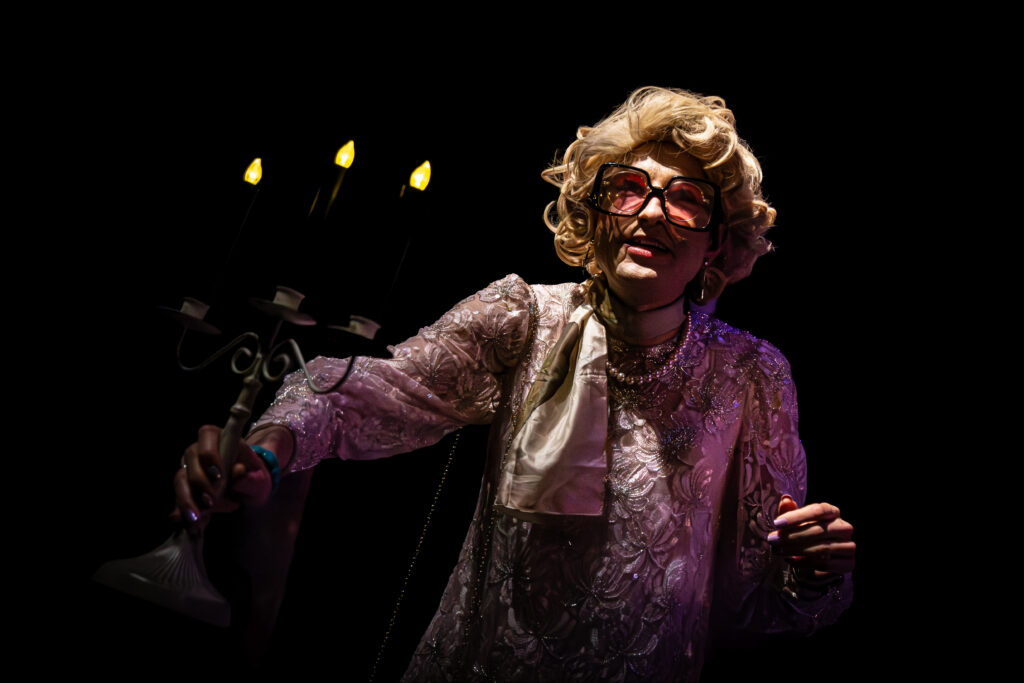 A performer dressed as an elderly woman with glasses holding a candelabrum with three lit candles during SpookyFest 2026 at The StageCrafts.