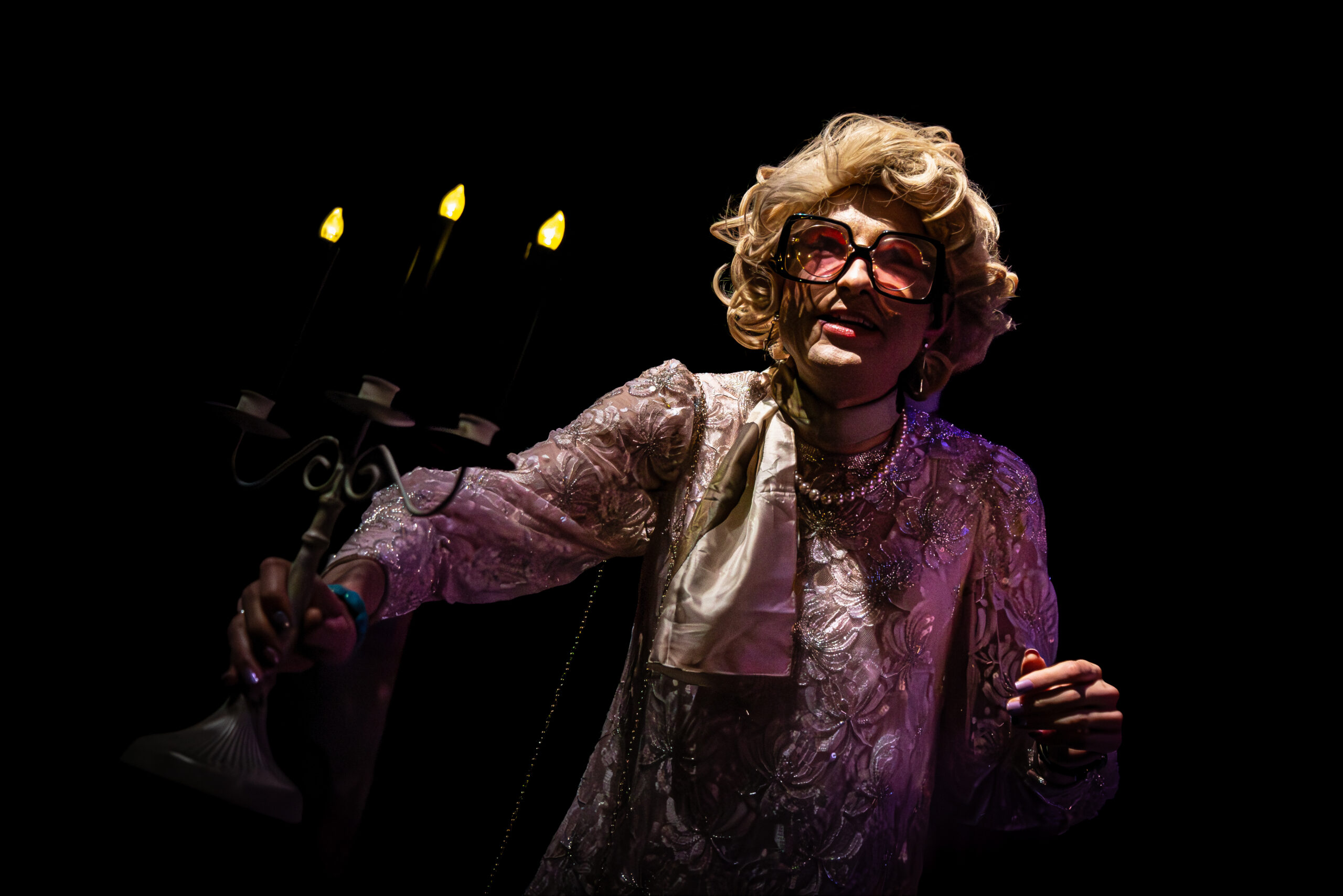 A performer dressed as an elderly woman with glasses holding a candelabrum with three lit candles during SpookyFest 2026 at The StageCrafts.