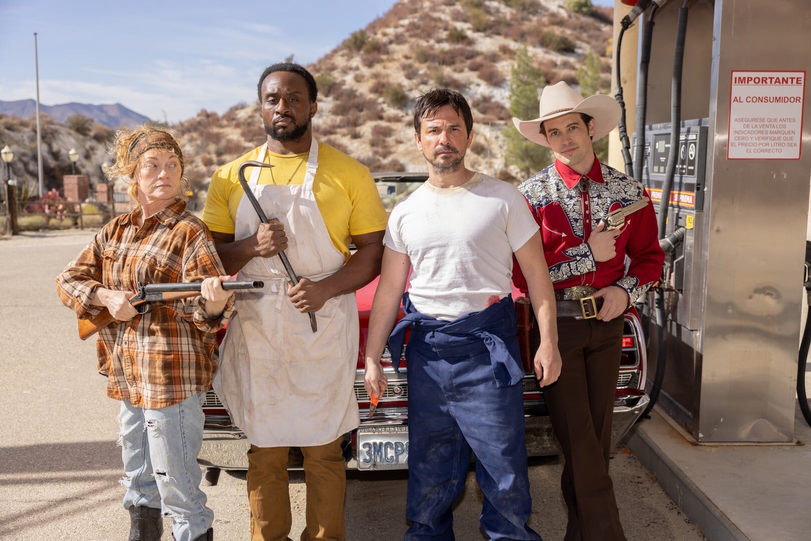 Group of four diverse actors in costume and casual wear standing outdoors in front of a car and a gas station, celebrating their success.