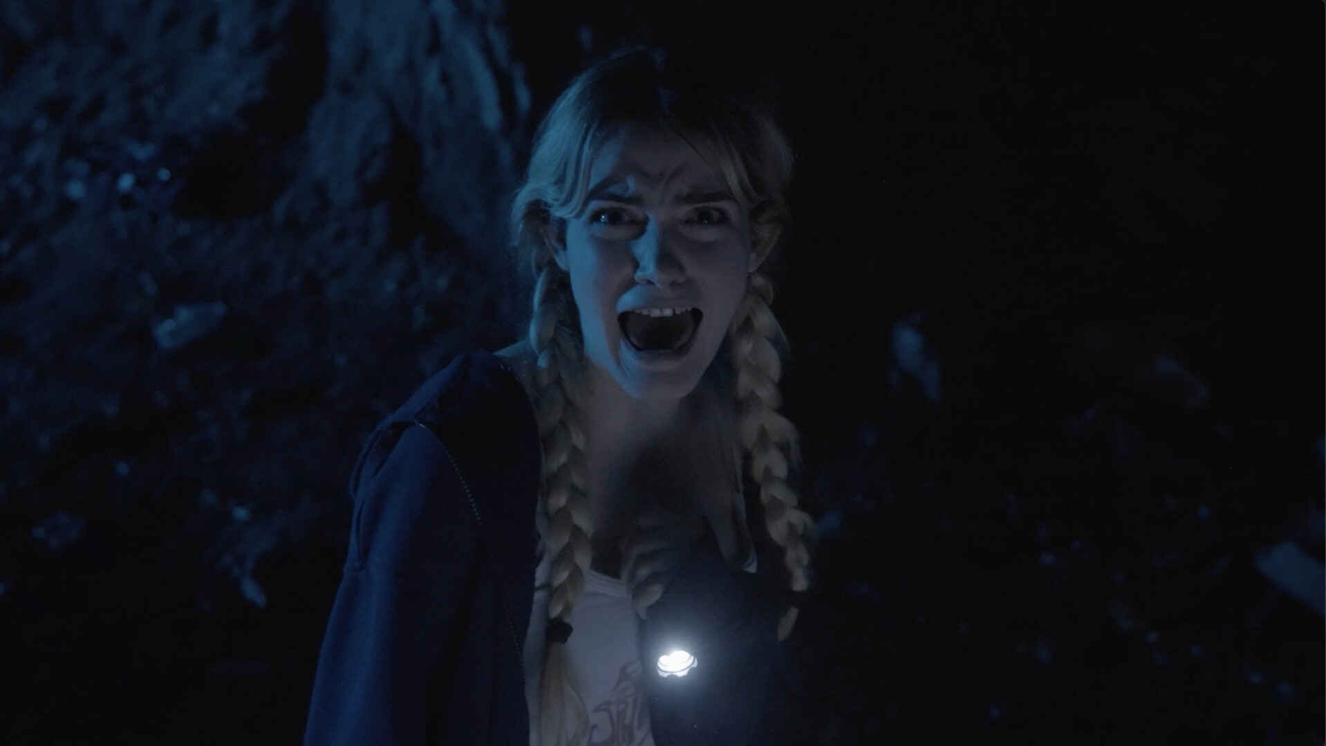 A young woman with braided hair and a flashlight in a dark, eerie setting, screaming with fear.