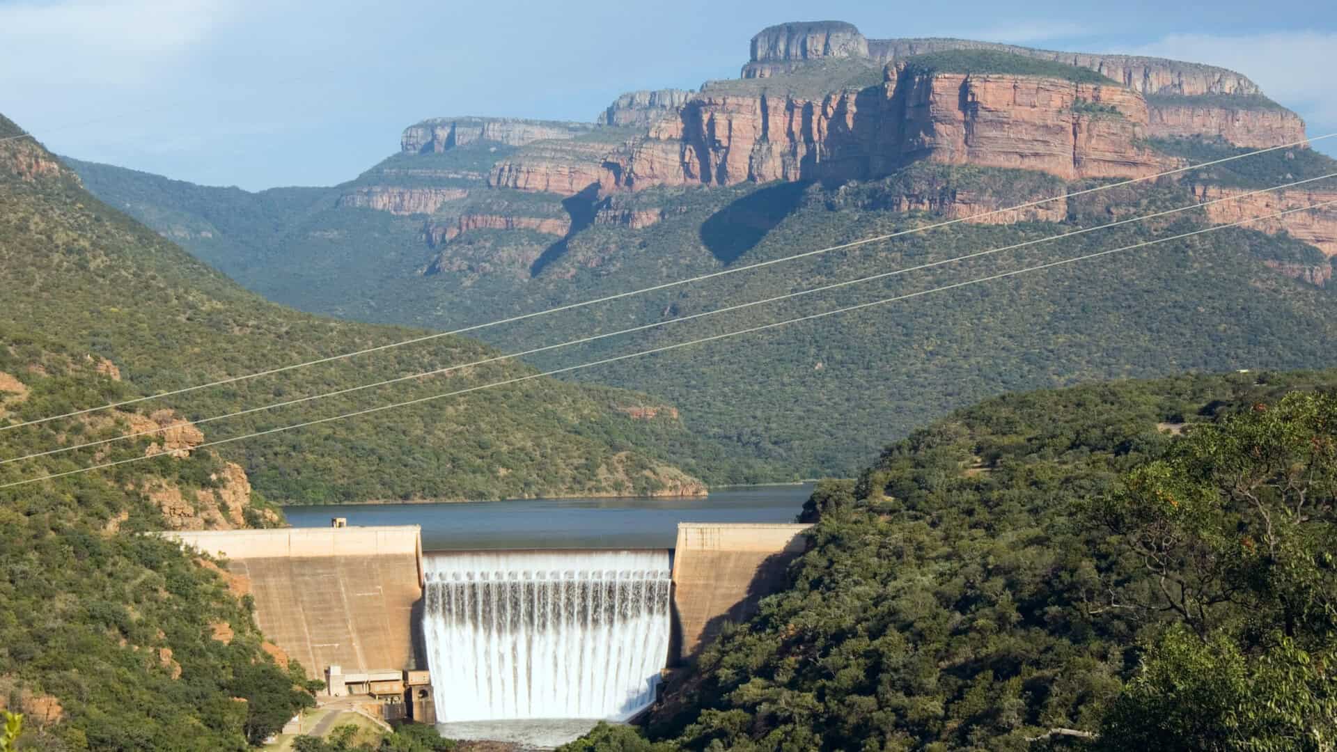 Scenic view of a dam with a waterfall, surrounded by lush green hills and rugged mountains in the background, under a clear blue sky in South Africa.