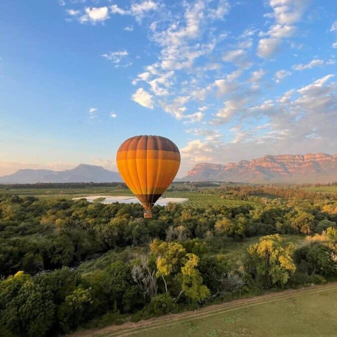 Hot air balloon floating over lush green landscape with mountains in the background during sunrise or sunset in South Africa.