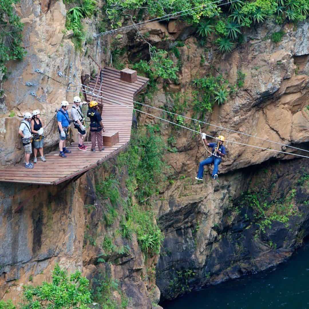 Adventure tourists crossing a suspension bridge and ziplining over a river in a lush, rocky canyon in South Africa.
