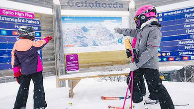 Kids and adult skiing enthusiasts pointing at a large ski map at a snowy ski resort, dressed in winter gear and helmets.