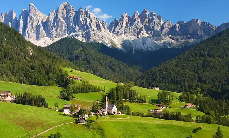 Vibrant green alpine landscape with a small village, church, and surrounding forests set against towering mountain peaks under a clear blue sky, perfect for winter and outdoor adventure enthusiasts.