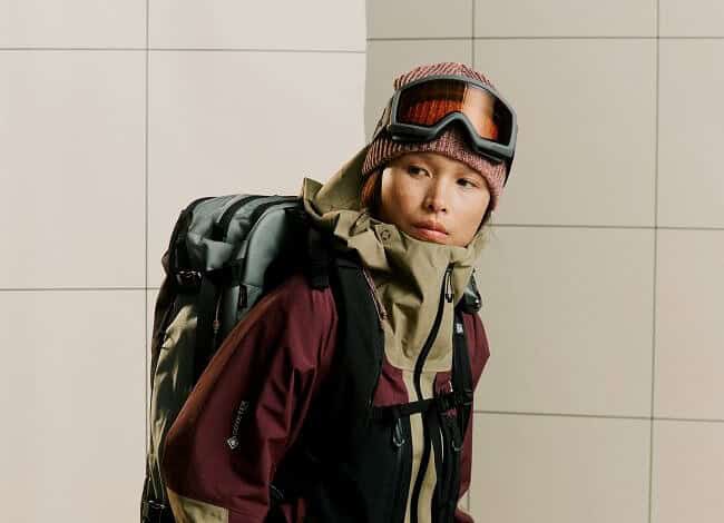 Backpacker in winter sports gear, wearing goggles and gloves, standing indoors, ready for winter adventure, showcasing premium winter clothing and equipment for snow and cold weather conditions.