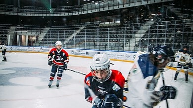 Young ice hockey players in action during a game at an indoor ice rink with bright overhead lights and spectators in the background.