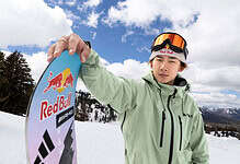 Young male snowboarder in the snow holding a snowboard with Red Bull logo, wearing winter gear and goggles, on a snowy mountain with trees and blue sky.