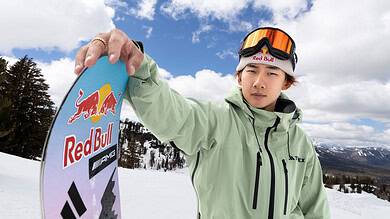 Young male snowboarder in the snow holding a snowboard with Red Bull logo, wearing winter gear and goggles, on a snowy mountain with trees and blue sky.