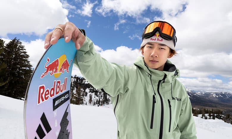 Young male snowboarder in the snow holding a snowboard with Red Bull logo, wearing winter gear and goggles, on a snowy mountain with trees and blue sky.