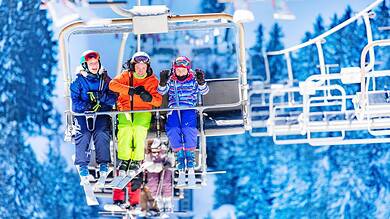 Family enjoying a ski lift ride at a winter resort with snowy slopes and pine trees in the background.