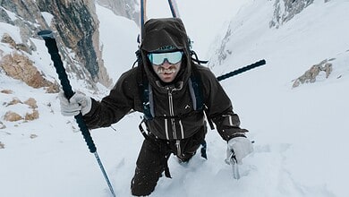 Backcountry skier ascending snowy mountain slope with ice axe and poles in winter landscape.