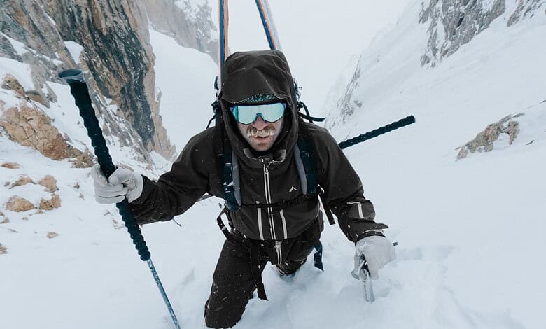 Backcountry skier ascending snowy mountain slope with ice axe and poles in winter landscape.