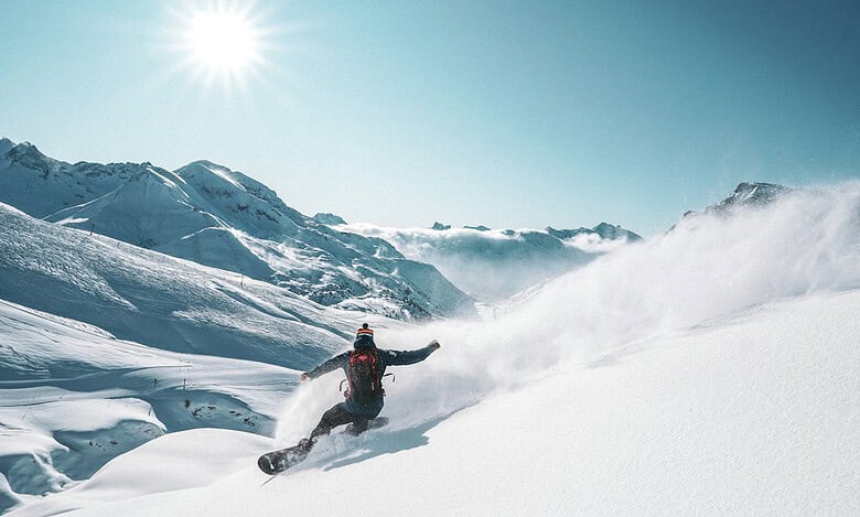 A snowboarder carving through fresh snow on a bright, sunny day in the mountains, with clear blue skies and snow-covered peaks in the background.