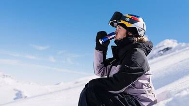 Female skier in winter gear drinking from a water bottle while sitting on snow-covered mountain slope, with clear blue sky and snowy peaks in the background.