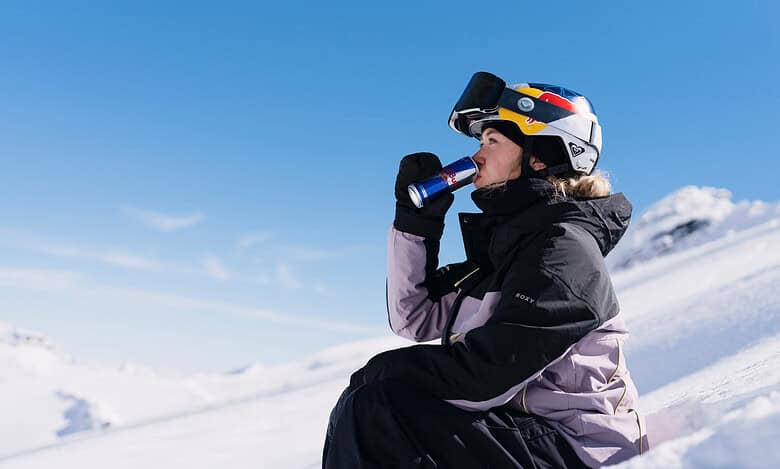 Female skier in winter gear drinking from a water bottle while sitting on snow-covered mountain slope, with clear blue sky and snowy peaks in the background.