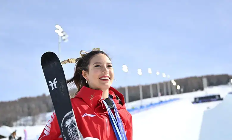 Young female freestyle skier in red jacket smiling with a snowboard at a snowy outdoor venue during daytime.