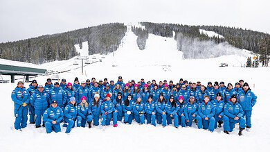 The Stifel U.S. Ski Team alpine athletes in blue gear pose together on snowy slopes with ski lifts and forested mountains in the background.