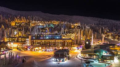 Nighttime view of a snowy mountain village illuminated with festive lights and decorated buildings. The scene captures the enchanting atmosphere of a winter holiday destination.