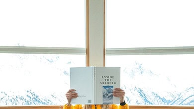 Person in a bright yellow jacket sits at a table by a large window, reading a book titled Inside the ARLBERG, with wine glasses and a dining setup on the round table.