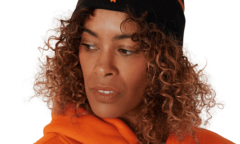 Woman wearing a black and orange knit beanie with a pom-pom and a bear mascot logo, paired with an orange hoodie.