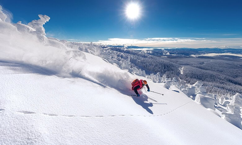 Skier in red gear descending a snowy slope under a bright sun in a winter landscape with clear blue sky and snow-covered trees.