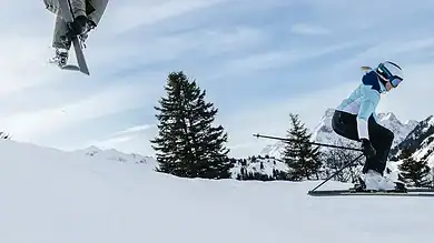 Two skiers performing jumps on snowy mountain slope with pine trees and cloudy sky in the background.