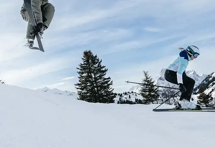 Two skiers performing jumps on snowy mountain slope with pine trees and cloudy sky in the background.