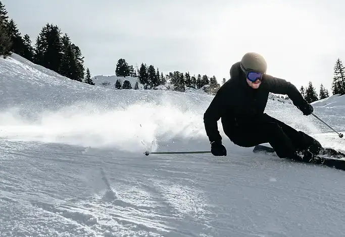 Skier in black gear carving through fresh snow on a sunny winter day with pine trees in the background.